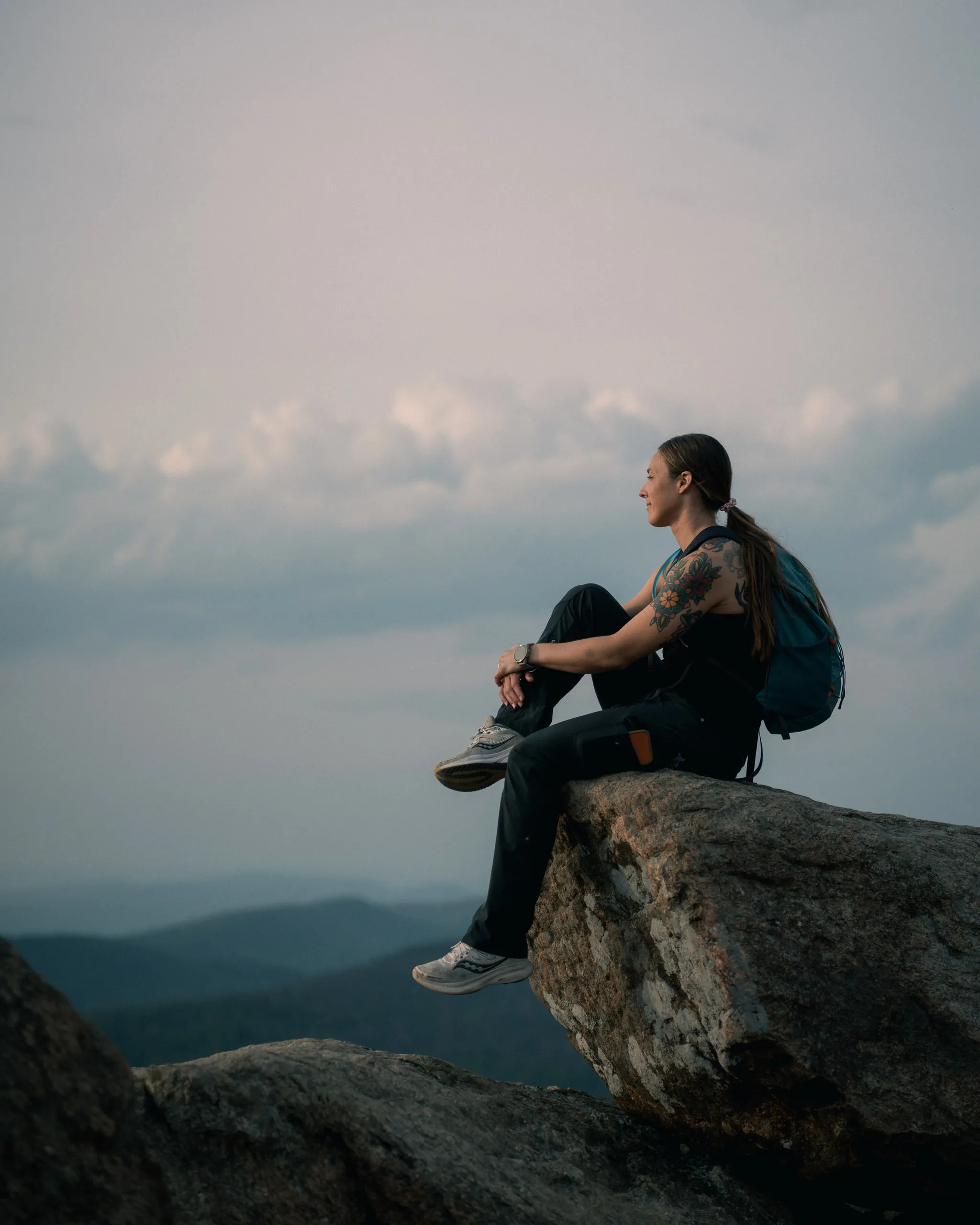 A hiker sits on a boulder at a ridge overlook, looking out over layered blue mountains